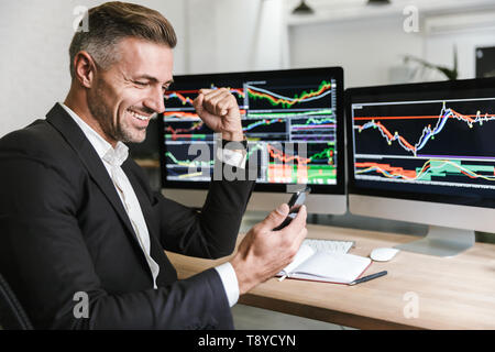 Portrait of male manager 30s wearing suit using mobile phone while working with digital graphics on computer in office Stock Photo