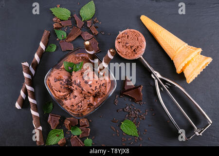 Scoops chocolate ice cream in glass bowl with wafer sticks, cone and chocolate on a black slate board. Focus on Bowl with scoops chocolate ice cream Stock Photo