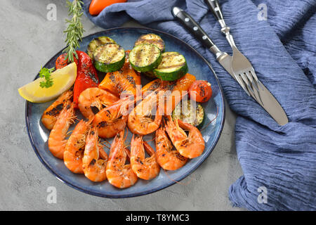 Grilled king prawns with mixed mediterranean vegetables served on a blue plate on a table with concrete look Stock Photo