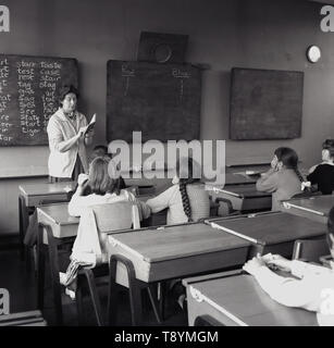 1960s, historical, children in a classroom , sitting at wooden desks, listening to the lady teacher, read from a book at the front of the class, England, UK. Stock Photo