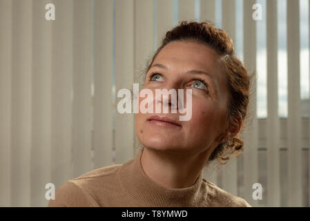 Close up indoor portrait of beautiful young 30s woman looking up and vertical blinds as background Stock Photo