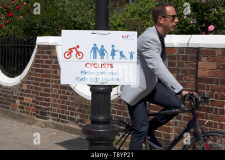 male cyclist passes a hammersmith and fulham council sign on the thames path asking cyclists to give way and cycle with courtesy, london, england Stock Photo