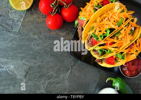 Hard shelled tacos with ground beef, lettuce, tomatoes and cheese. Top view, corner border on a dark background with copy space. Stock Photo