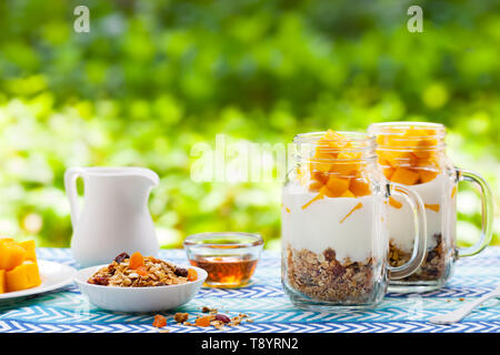 Healthy breakfast, dessert. Fresh mango fruit with yogurt and granola in jars. Outdoor background. Copy space. Stock Photo