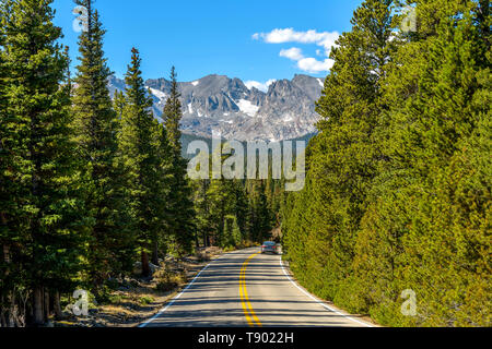 Mountain Road - A mountain road winding through in a dense evergreen forest, with Indian Peaks and Glaciers rising high in background. Colorado, USA. Stock Photo