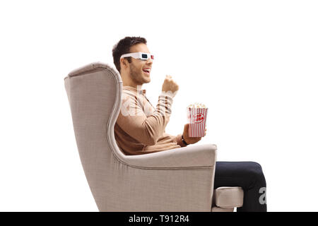 Young man in an armchair wearing a pair of 3D glasses and eating popcorn isolated on white background Stock Photo