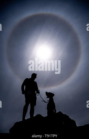 Aberystwyth Wales UK, Thursday 16 May 2019  UK Weather: A man and his dog arr silhouetted as perfect circular sun halo , formed as the rays of the sun shine through ice crystals in the upper atmosphere,  fills the sky above Aberystwyth. The halos, always at 22º from the sun,  often  indicate that rain will fall within the next 24 hours, since the cirrostratus clouds that cause them can signify an approaching frontal system. The  weather is set to change overnight from the fine warm conditions to cooler and wetter days, more typical of mid May.  photo credit Keith Morris / Alamy Live News Stock Photo