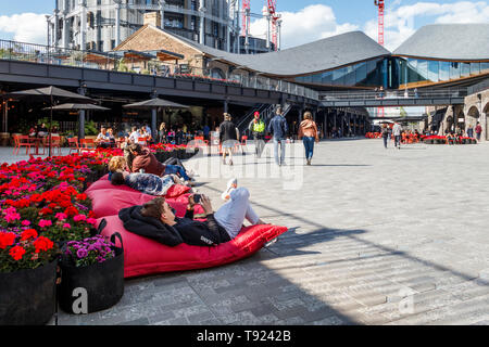 People lounging on red cushions by a bed of bright red flowers in the recently opened public space at Coal Drops Yard, King's Cross, London, UK, 2019 Stock Photo