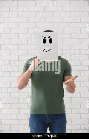 Young man hiding face behind sheet of paper with drawn emoticon against white brick wall Stock Photo