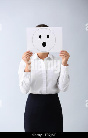 Young woman hiding face behind sheet of paper with drawn emoticon on light background Stock Photo