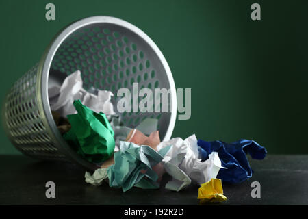 Overturned recycle bin with crumpled papers on table against color background Stock Photo