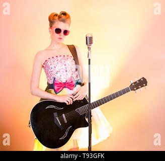 Stylish girl guitarist singer in pink glasses with black guitar, vintage microphone. Teenager musician in funny vintage retro dress on colored rainbow Stock Photo