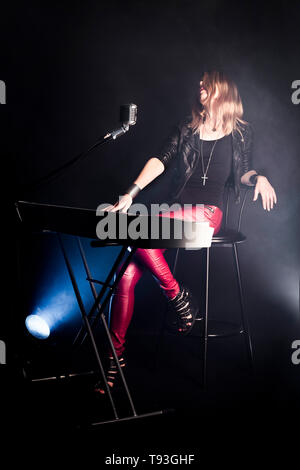 Blonde girl musician pianist plays perform on white digital piano, sits on chair, sings into retro microphone. Woman teacher shows how to play music Stock Photo