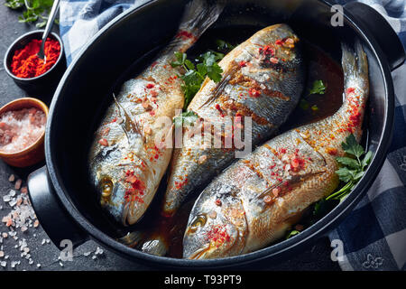 three Raw dorado fish marinated with spices, sea salt, ginger, soy sauce and herbs in a baking dish on a concrete table with ingredients, view from ab Stock Photo