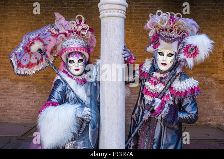 Portraits of a masked couple in beautiful creative costumes, posing in a backyard with pillars, celebrating the Venetian Carnival Stock Photo