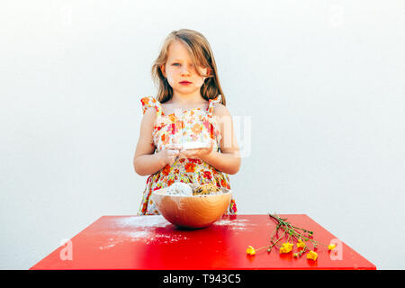 portrait of a little girl with a soiled face flour, isolated on white background, wearing a flower theme dress. A bowl with cookies and yellow flowers Stock Photo