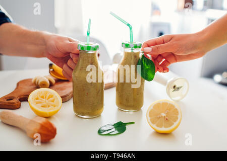 Bottles with spinach and banana smoothie with ingredients on kitchen table. People holding drinks. Healthy detox diet Stock Photo