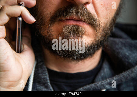 Close up portrait of bearded man talking on smart phone, focus on lips, no eyes, secret talk, criminal threating someone Stock Photo