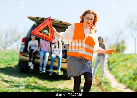 A woman in orange vest set up breakdown triangle stands near a broken car - of road Stock Photo