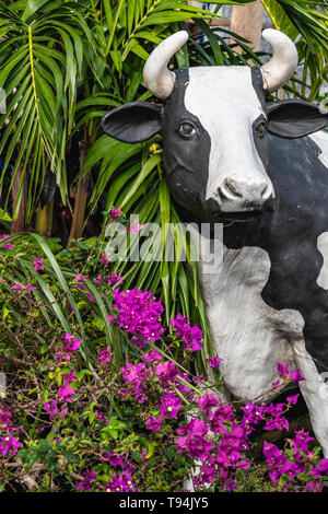 black and white cow statue and bright pink bougainvillea with green palm leaves behind Stock Photo