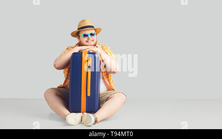 Fat funny man with a suitcase smiling on a gray background. Stock Photo