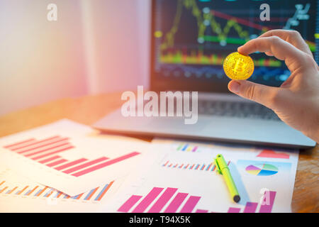 Hand holding bitcoin in front of laptop and diagrams on desk Stock Photo