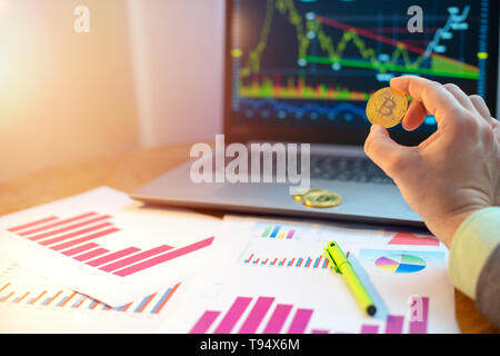 Hand holding bitcoin in front of laptop and diagrams on desk Stock Photo