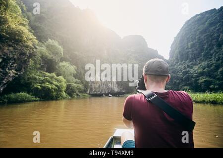 Photographer with camera on boat. Young man taking photo of river against karst formation near Tam Coc in Ninh Binh province, Vietnam. Stock Photo