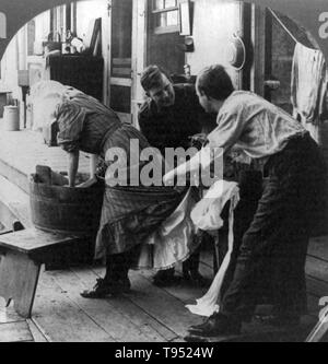 Entitled: "Wringer up George, quick!" show a woman doing laundry in tub, as two men put bottom of her dress and skirt through wringer. Cropped stereograph photographed by E.W. Kelley, 1906. Stock Photo