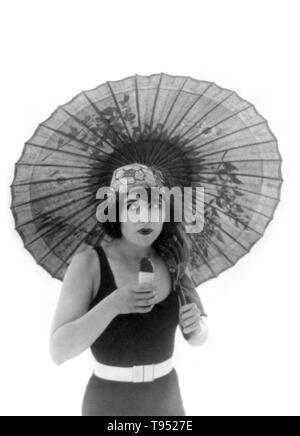 Entitled: 'Betty Compson with umbrella, in bathing suit, facing slightly right, eating Eskimo Pie.' Betty Compson (March 19, 1897 - April 18, 1974) was an American actress and film producer. Most famous in silent films and early talkies, she is best known in her performances in The Docks of New York and The Barker, the latter earning a nomination for the Academy Award for Best Actress. Her popularity allowed her to have creative control over her films as she was also able to produce. Stock Photo