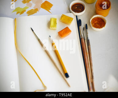 On a light table are art supplies: paints, pencil, brushes, paper palette and an empty notebook for drawing. Everything is prepared to create an illus Stock Photo