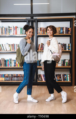 Two cheerful young girls students studying at library, carrying backpacks Stock Photo
