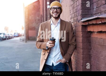 Handsome young man in casual wear holding disposable cup and smiling while walking through the city street. Stock Photo