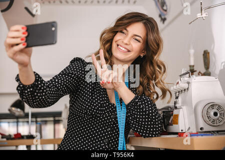 Smiling young woman seamstress working at the workshop, using sewing machine, taking a selfie Stock Photo