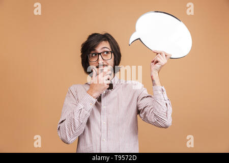 Portrait of clever man 20s wearing eyeglasses and bow tie holding blanc idea bubble while standing isolated over beige background in studio Stock Photo