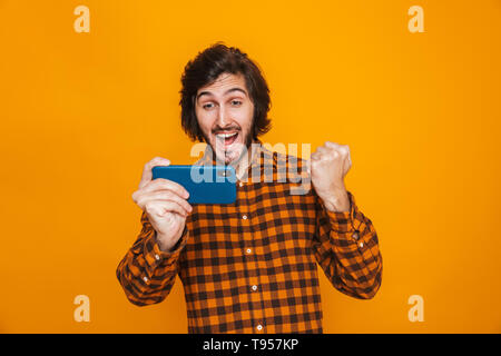 Image of joyous man wearing plaid shirt playing video games on smartphone while standing isolated over yellow background in studio Stock Photo