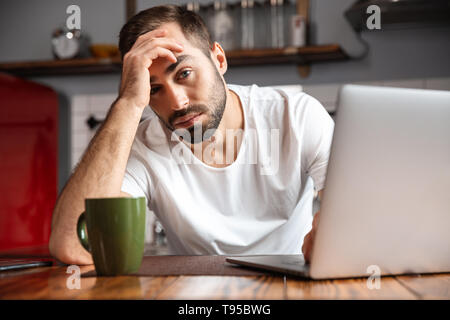 Photo of displeased man 30s wearing casual t-shirt using silver laptop while sitting at table in modern apartment Stock Photo