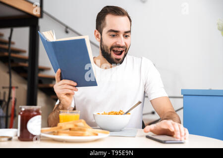 Handsome young man having breakfast while sitting at the kitchen, reading a book Stock Photo