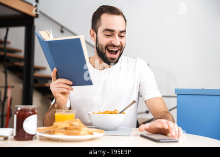 Handsome young man having breakfast while sitting at the kitchen, reading a book Stock Photo