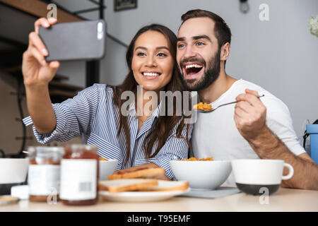 Happy multiethnic couple having breakfast at the kitchen, taking a selfie Stock Photo
