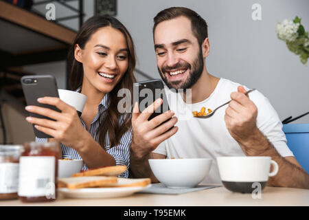 Happy multiethnic couple having breakfast at the kitchen, using mobile phones Stock Photo