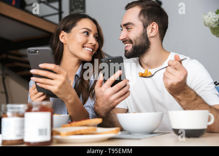Happy multiethnic couple having breakfast at the kitchen, using mobile phones Stock Photo