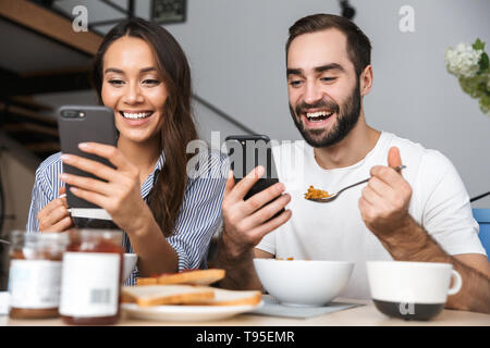 Happy multiethnic couple having breakfast at the kitchen, using mobile phones Stock Photo