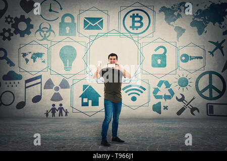 Guy presenting a trendy mobile phone recommending a new device showing thumb up gesture and 3D digital icons on the background. Media technology moder Stock Photo