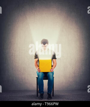 Curious man sitting on a chair in a dark room holding yellow box on knees, looking inside as a magic light come out of the bin. Surprise concept, rece Stock Photo