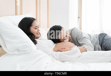 Asian Handsome man is listening to his Asian beautiful pregnant wife's tummy and smiling. Family love concept. Stock Photo