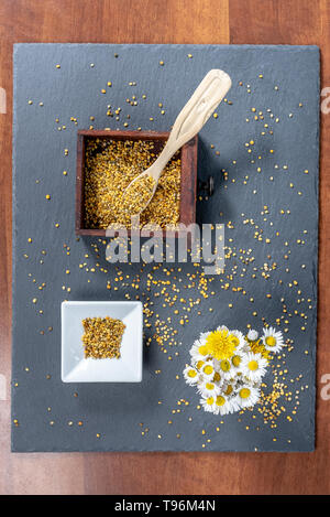 photo composed of a square bowl of white ceramic, a wooden box full of pollen grains of bees and a bunch of daisies, on a slate board, top view Stock Photo