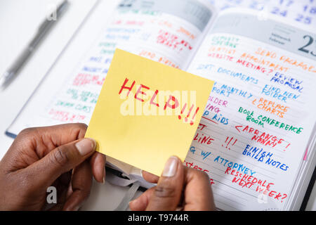 Close-up Of Person's Hand Holding Adhesive Note With Help Text Over Diary Stock Photo