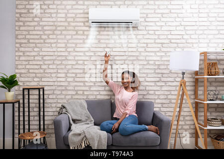 Young African Woman Switching On Air Conditioner With Remote Control At Home Stock Photo