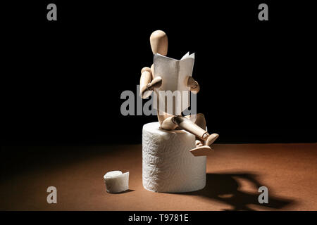 Small wooden mannequin reading newspaper while sitting on toilet paper roll against dark background Stock Photo